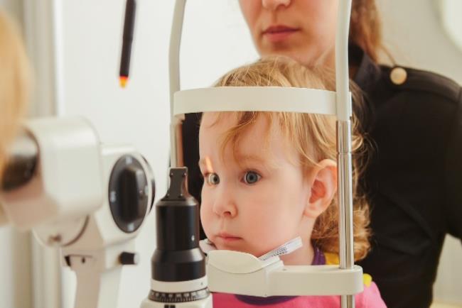 ילדה קטנה על ברכי אמה עוברת בדיקת ראייה באמצעות רטינוסקופ לאבחון של פזילה 